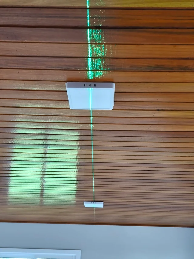 Linha de laser atravessando luminárias em um forro de madeira durante a instalação.